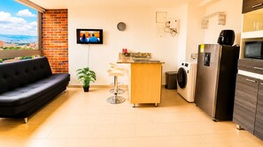 Ein kompakter Wohnbereich mit einer Couch, einer kleinen Küchenzeile inklusive Waschmaschine und einem an der Wand befestigten Fernseher.