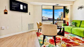 A modern living room with a green sofa, a beige armchair, a small table, and a large window with a city view.