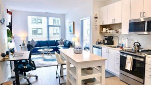 Et moderne køkken med hvide skabe, en mikrobølgeovn og komfur. Der er et spiseområde med et bord og stole, og en blå sofa i baggrunden.