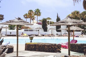 A poolside area with lounge chairs and umbrellas, a stone wall, and a clear view of the pool. - Apto 115 (Costa Teguise)