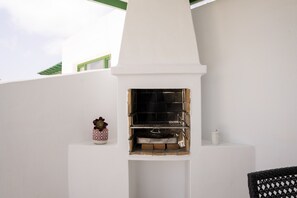 A white outdoor fireplace with a brick interior, a small decorative pot, and a white container on the right side. - Apto 115 (Costa Teguise)