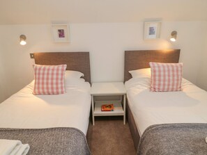 A twin room with two beds, each with a checkered pillow, a nightstand with a book, and wall-mounted lights. - Harvest Cottage 1 (Berwick-upon-Tweed)