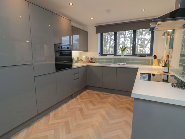 Cozinha moderna com armários cinza, forno embutido, piso de madeira em padrão herringbone, janela com vista para árvores e bancada com uma garrafa de vinho.