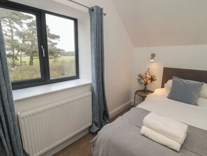 Quarto com uma janela grande, radiador, mesa de cabeceira com abajur, cama com almofadas e vista para árvores lá fora.