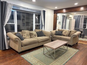 A living room with two sofas, a coffee table, a patterned rug, and a dining area in the background.