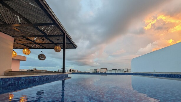Una piscina infinita con mosaicos azules, un área de estar techada con lámparas de mimbre y un edificio moderno al fondo.