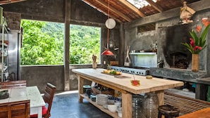 A rustic kitchen with a large island, a stone fireplace, and a view of lush greenery outside. - Vereda Yelapa : Abiu (Yelapa)