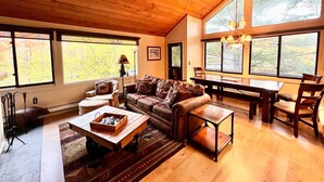Ein geräumiges Wohnzimmer mit Holzbalkendecke, großen Fenstern, einer Couch, einem Couchtisch und einem Essbereich.