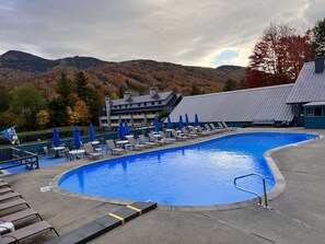A swimming pool with lounge chairs and umbrellas, a building with a sloped roof, and a mountainous backdrop.