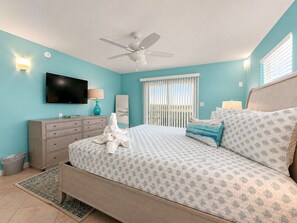 Ein Schlafzimmer mit Bett, Deckenventilator, Fernseher, Kommode und Fenster mit Jalousien.