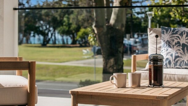 Ein Holztisch mit Kaffeemaschine und zwei Tassen, ein Fenster mit Blick auf Bäume und eine Wiese, sowie ein Stuhl mit Sitzkissen.
