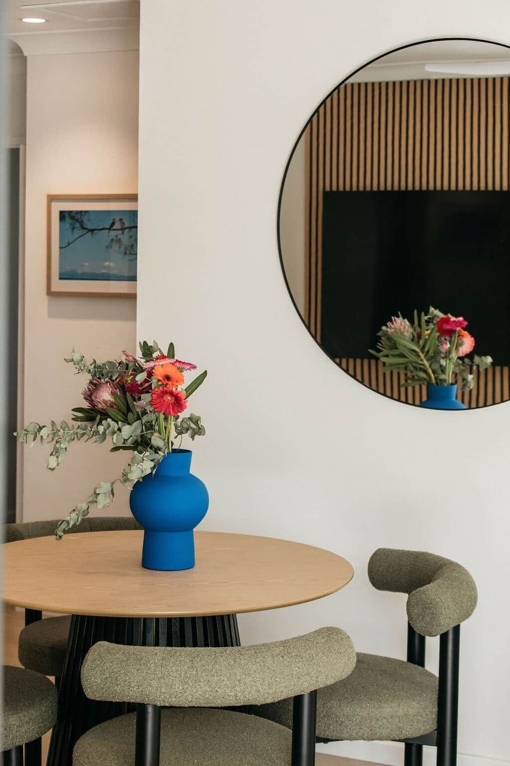 Ein runder Tisch mit einer blauen Blumenvase, grün gepolsterten Stühlen und einem runden Spiegel an der Wand.