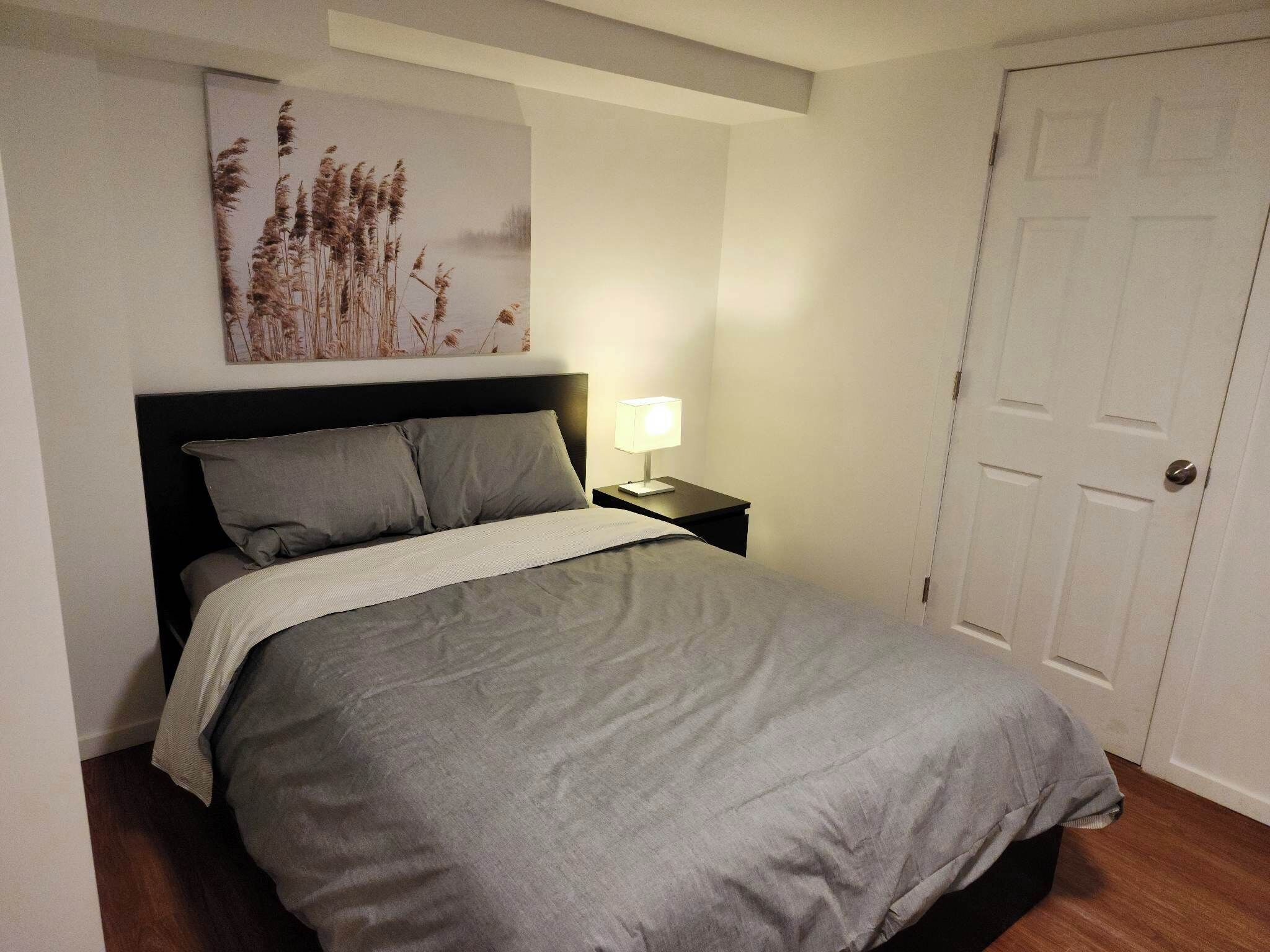 Une chambre à coucher avec un lit, une table de chevet avec une lampe, une porte et un tableau accroché au mur.
