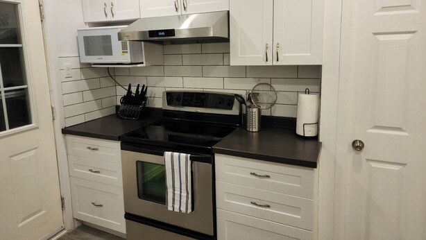 Una cocina con gabinetes blancos, una campana extractora de acero inoxidable, un microondas, una estufa y un lavamanos.
