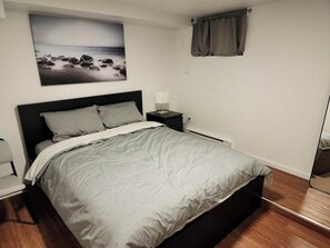 A bed with a grey bedspread, a nightstand with a lamp, a wall-mounted painting of a beach, and a window with curtains.