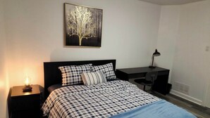 Un dormitorio con cama, escritorio, silla, mesita de noche, lámpara y un cuadro colgado en la pared.