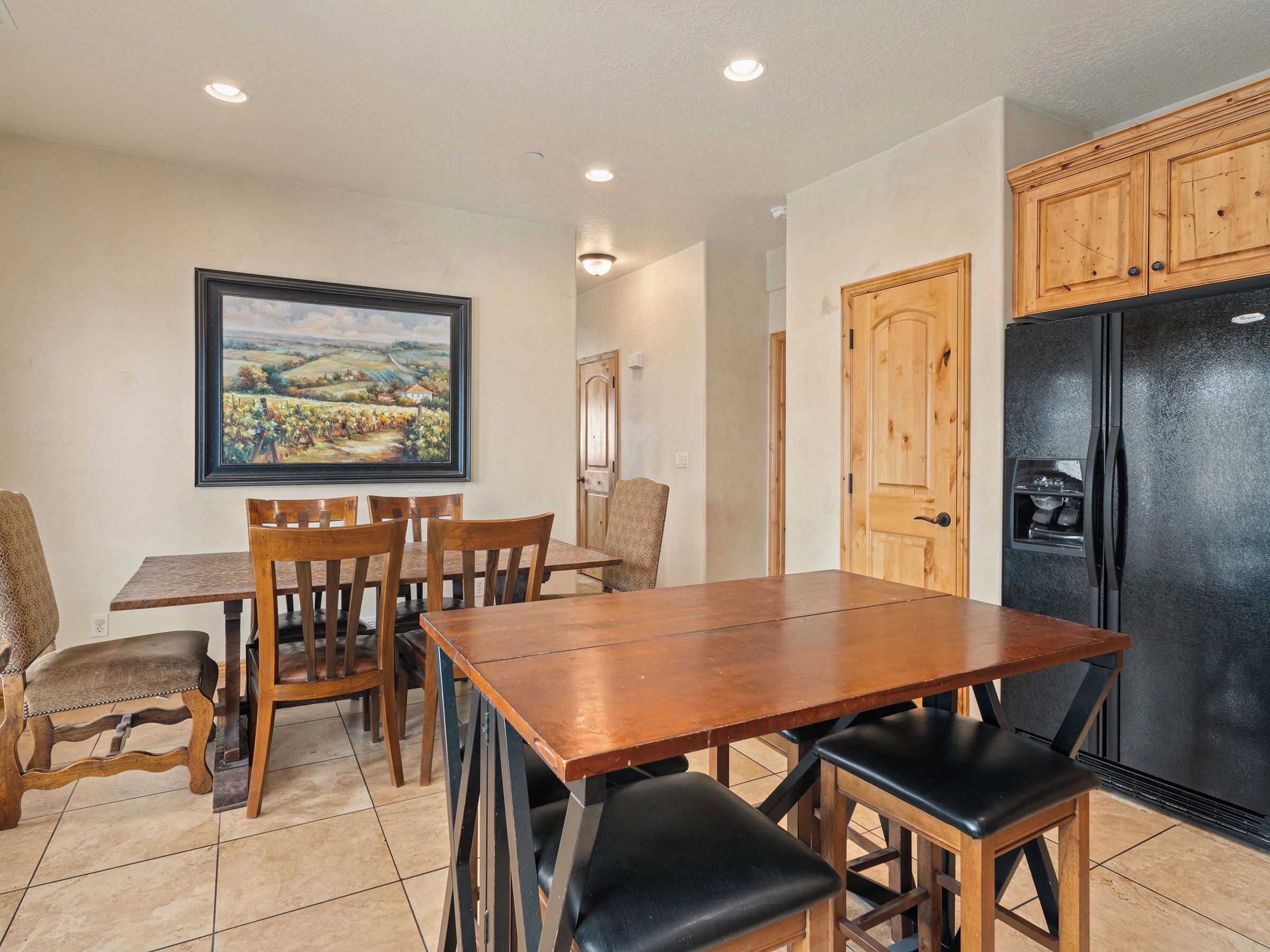 A dining area with a wooden table and chairs, a black refrigerator, wooden cabinets, and a framed landscape painting on the wall.