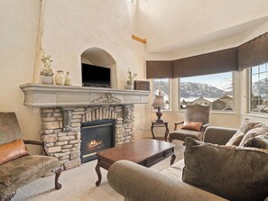 A living room with a stone fireplace, a television mounted above it, and a view of snow-capped mountains through large windows.