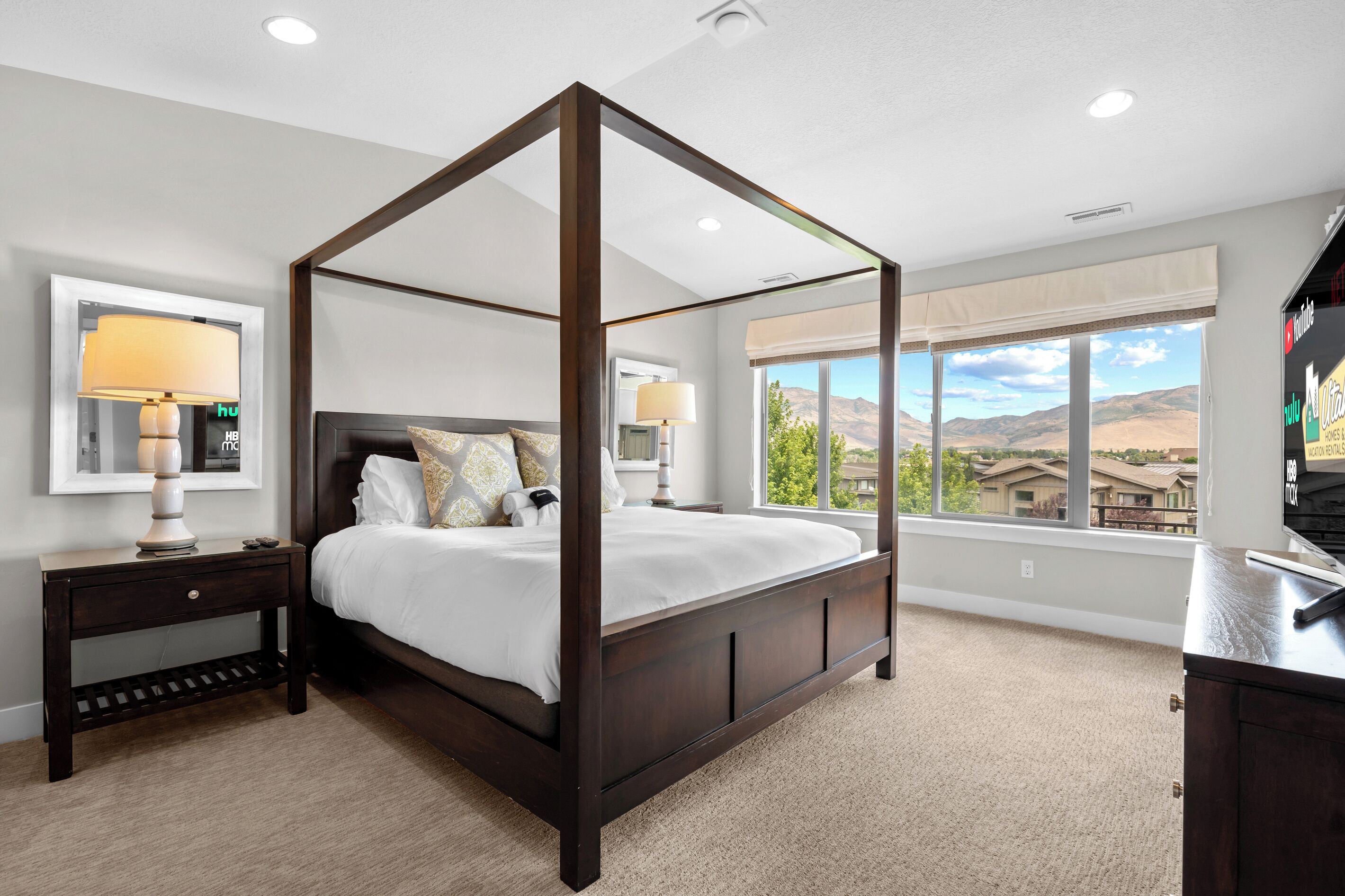 A four-poster bed with a dark finish, a bedside table with a lamp, and a large window offering a view of a mountainous landscape.