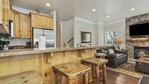 A kitchen with wooden cabinets, a stainless steel refrigerator, a stone fireplace, and a bar area with stools. - Lakeside Getaway LS14A | Hot Tub | Dog-Friendly (Huntsville)