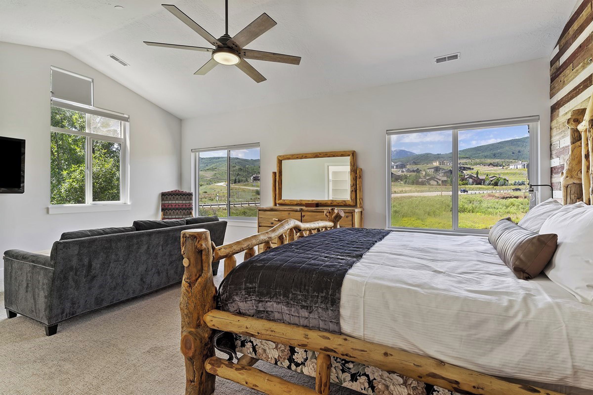 Une chambre à coucher avec un grand lit, une tête de lit en bois, un ventilateur de plafond et une vue sur la campagne, visible à travers de grandes fenêtres.