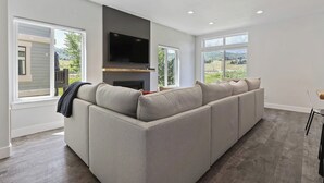 Un salon moderne avec un canapé d’angle, un téléviseur à écran plat et de grandes fenêtres donnant sur l’extérieur.