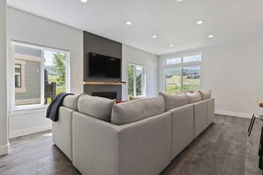 A modern living room with a sectional sofa, a flat-screen TV, and large windows offering a view of the outdoors. - Pineview Lake Retreat LS22 | Hot Tub | Pool | Desk (Huntsville)