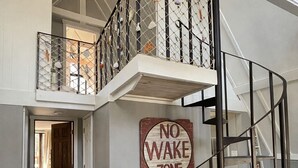 Ein geräumiger Innenraum mit Holzboden, einem 'No Wake Zone'-Schild, einer Wendeltreppe und einem Balkon mit Metallgeländer.