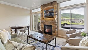 Una sala de estar con chimenea de piedra, comedor y vistas a un edificio y colinas.