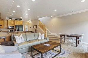A living room with a sofa, coffee table, and dining table with chairs. There is a kitchen area with cabinets and appliances. A staircase is visible in the background. - Lakeside Ski Den LS40 |Game Room | Pool | Hot Tub (Huntsville)