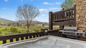 A hot tub with a stone wall and a grill, overlooking a green landscape with buildings. - Paddle & Ski Retreat LS43A |Pool |Hot Tub|Pineview (Huntsville)