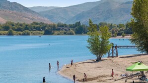 Ein Strand am Seeufer mit badenden und spazierenden Personen, einer Anlegestelle und einem grünen Sonnenschirm.