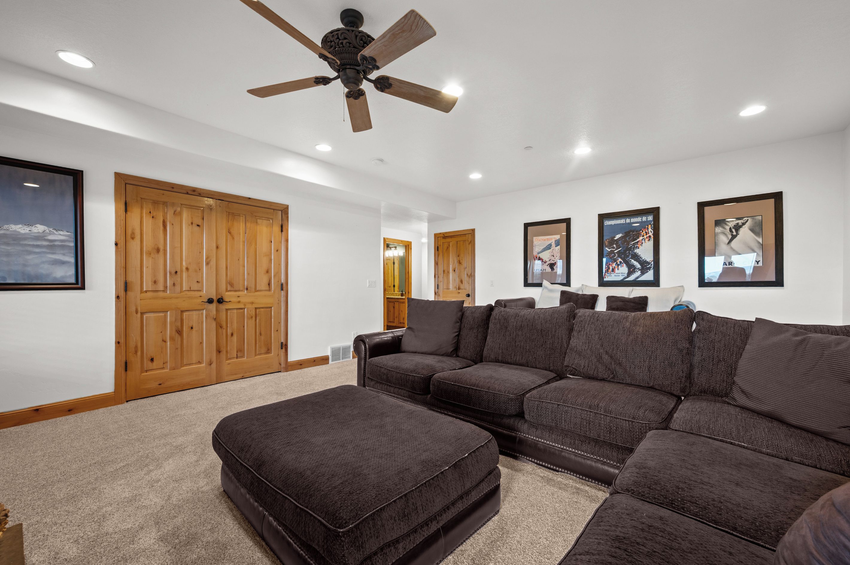 A living room with a sectional sofa, ottoman, wooden doors, ceiling fan, and wall-mounted artwork.