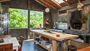 A rustic kitchen with a large island, a stone fireplace, and a view of lush greenery outside. - Vereda Yelapa: Passion Fruit (Yelapa)