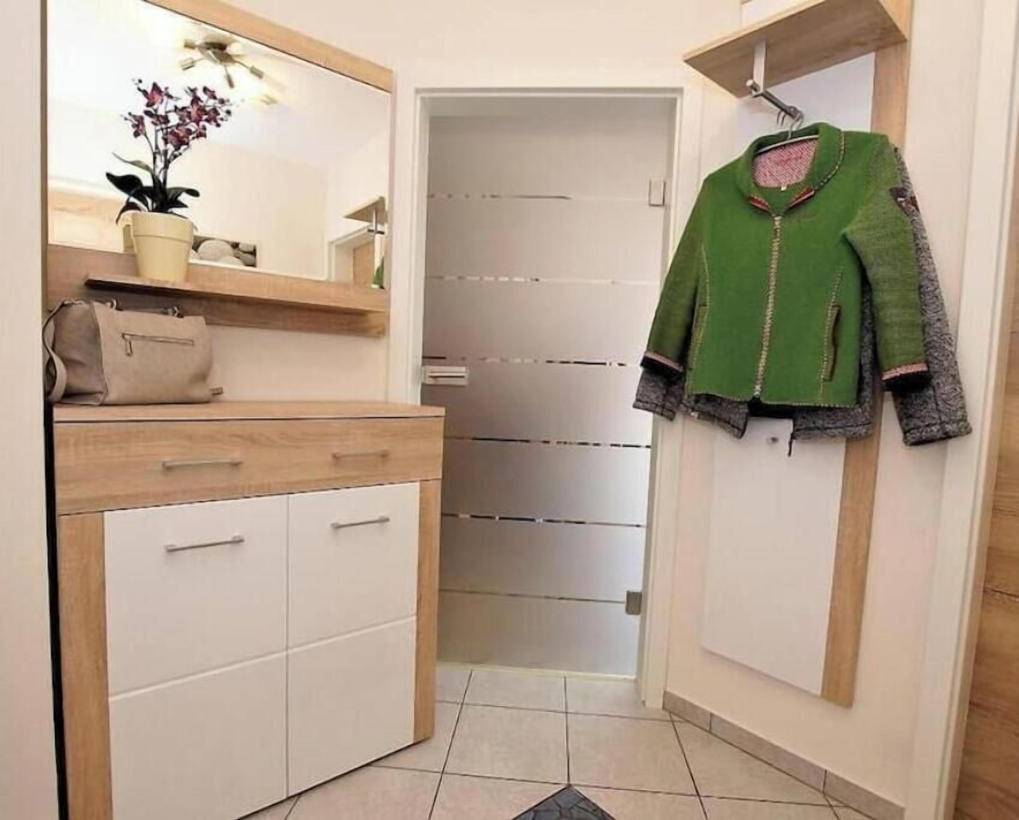 Een hal met een spiegel, een potplant, een beige handtas en een groen jasje aan de muur.