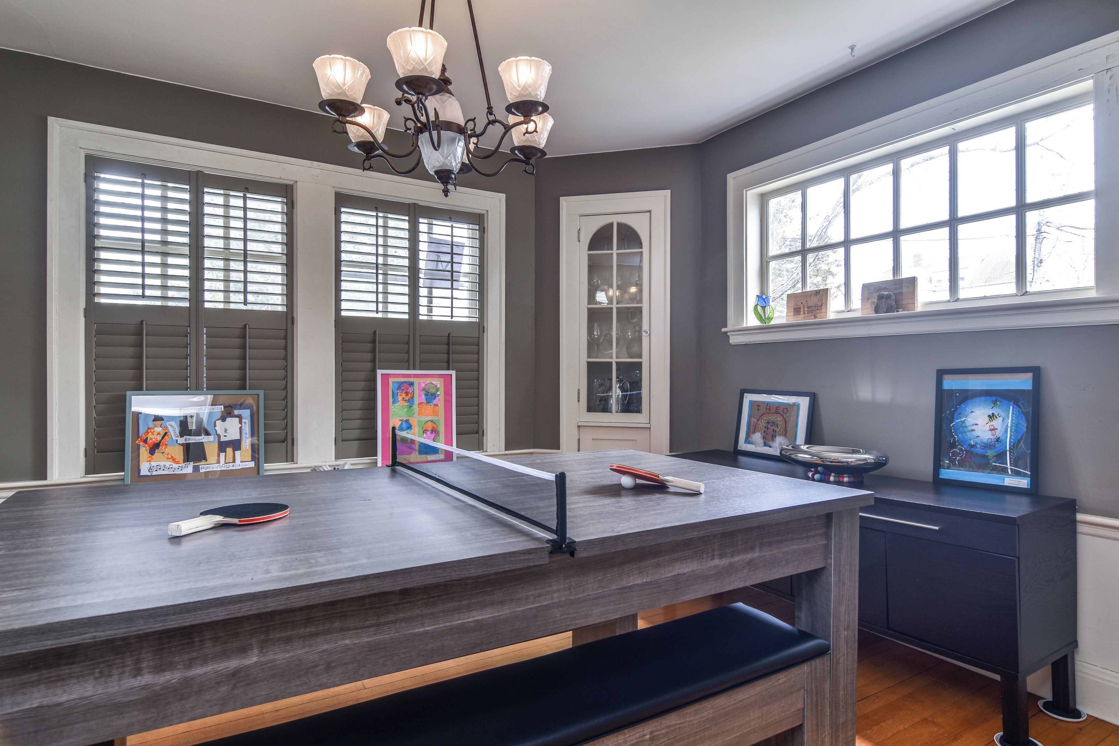 A room with a ping pong table, a chandelier, a window with blinds, and artwork on the wall.