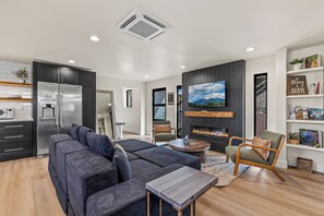 A modern living room with a sectional sofa, a fireplace, a flat-screen TV, and a kitchen with a refrigerator. - Epic Rooftop Views - Luxury DT FLAG - 2 King Beds (Flagstaff)