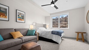 Ein Schlafzimmer mit einem Bett, einem grauen Sofa, einem braunen Hocker, einem Nachttisch mit Lampen, einem Deckenventilator und einem Fenster mit Blick auf Felsen.