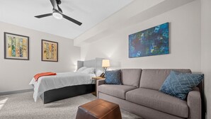 A modern hotel room with a bed, sofa, ottoman, ceiling fan, artwork, and a lamp. - Water's Edge 102C | Bear Lake| Lakeside Unit (Garden City)