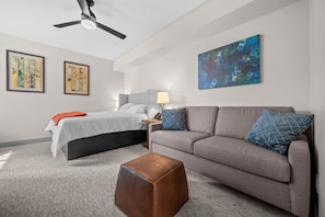 A modern hotel room with a bed, sofa, ottoman, ceiling fan, artwork, and a lamp. - Water's Edge 102C | Bear Lake| Lakeside Unit (Garden City)