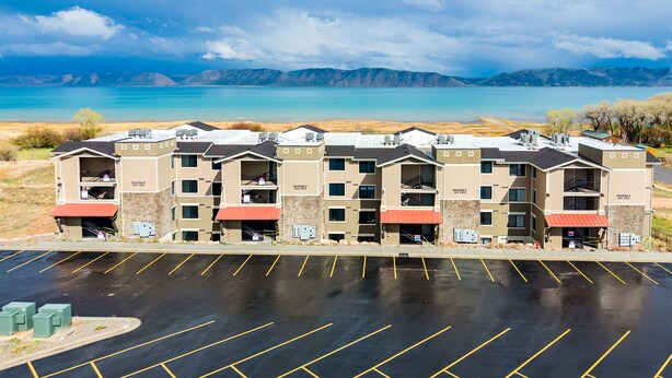 A multi-story hotel with a parking lot in front, surrounded by mountains and a lake. - Water's Edge 102C | Bear Lake| Lakeside Unit (Garden City)