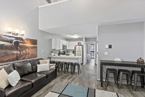 Ein modernes Wohnzimmer mit einer Ledersofa, einem Couchtisch und einem großen Gemälde. Es gibt eine Küchenzeile mit weißen Schränken und eine Bar mit Hockern.
