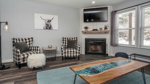 Un salon avec une cheminée, des fauteuils à carreaux, une table basse et un téléviseur fixé au mur.