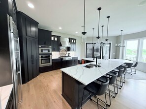 A modern kitchen with dark cabinetry, stainless steel appliances, a large island with bar stools, and pendant lighting. - Newly Remodeled, w/ Hot Tub - Lake Views spacious (Wellsville)