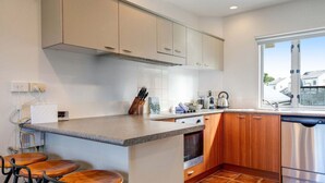 A kitchen with a central island, bar stools, wooden cabinets, stainless steel appliances, and a window with a view. - Henderson Retreat 3BR Townhouse with Parking (Auckland West)