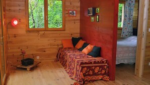 Una cabaña de madera con una cama, un banco con cojines, un ventanal con vistas a árboles y una pequeña mesa con una flor.