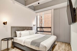 Ein modernes Schlafzimmer mit einem großen Bett, einem Nachttisch, einem an der Wand montierten Fernseher und einem Fenster mit Blick auf ein Backsteingebäude.