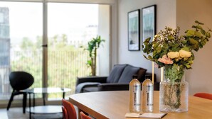 Un salon moderne avec une table en bois, deux bouteilles, un vase de fleurs, un canapé et une vue sur la verdure à l’extérieur.