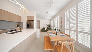 A modern kitchen with a central island, a dining area with a wooden table, and a large window with blinds. - Sage Palmview - Home Away From Home (Mooloolaba Region)