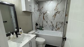 A modern bathroom with a white sink, toilet, and bathtub, marble tiles, and a mirror with lights. - The Urban Retreat (Calgary)